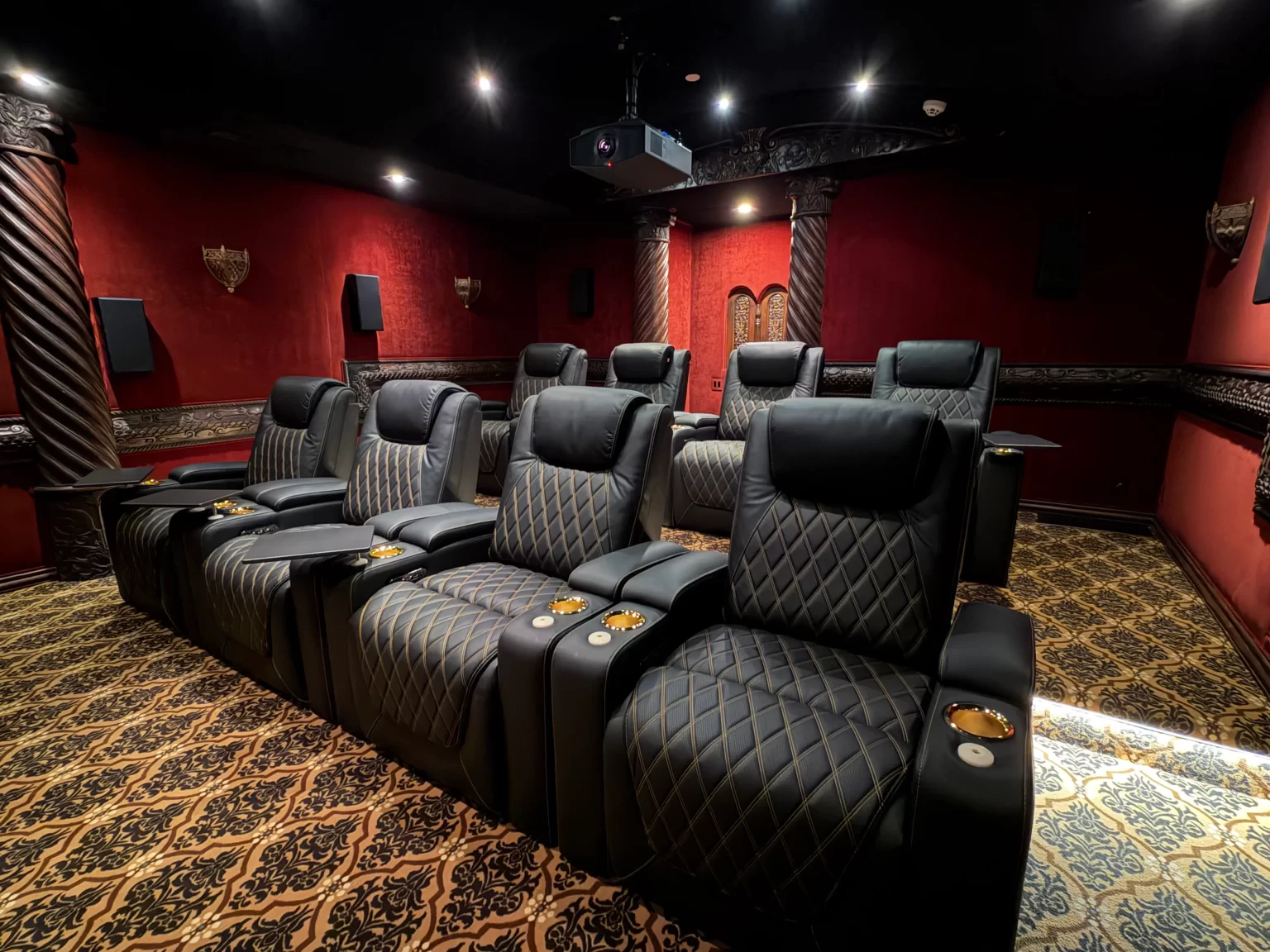 Two rows of black theater recliners beneath a ceiling-mounted projector in a red dedicated cinema